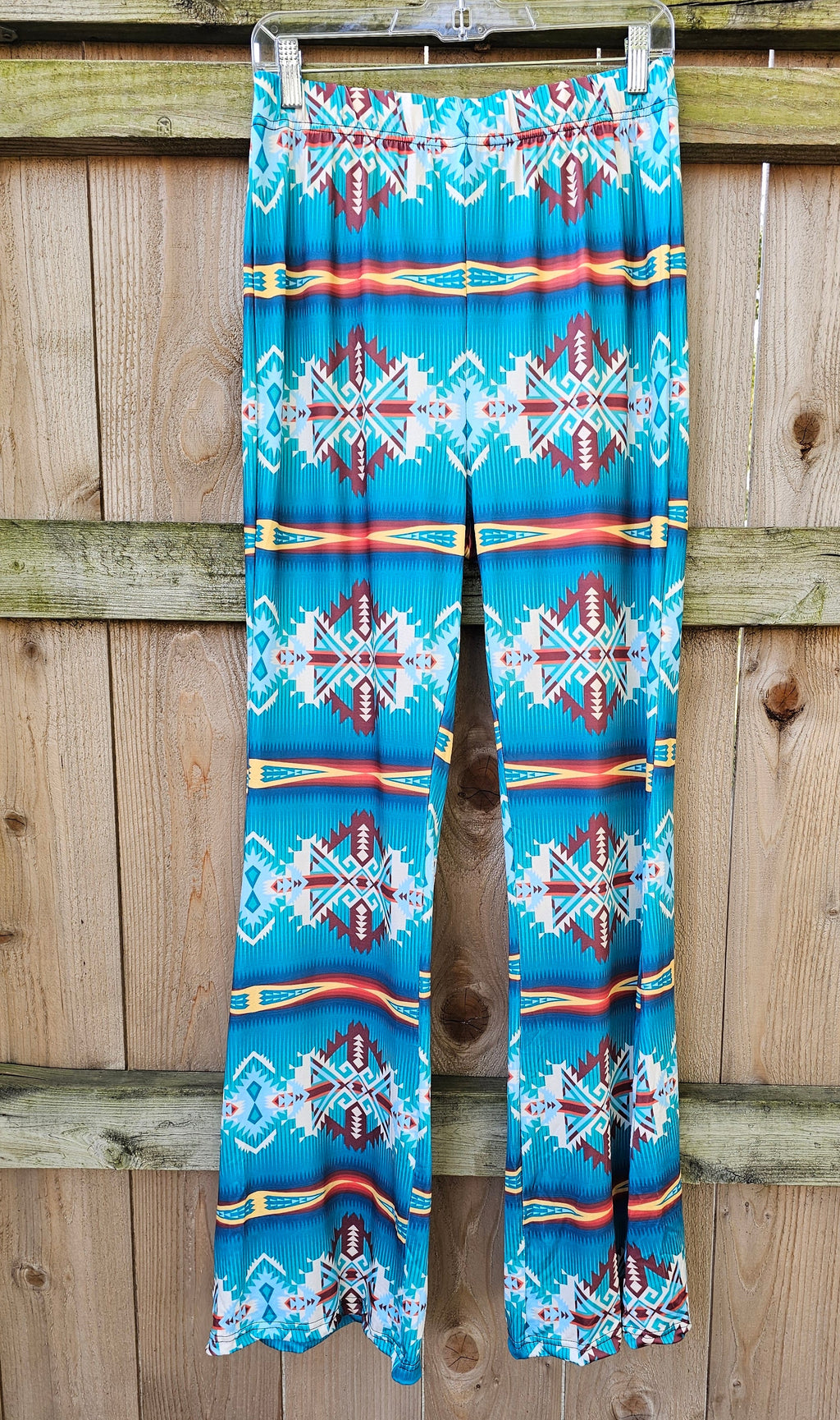 Teal Aztec Flare Pants hanging on a wooden fence, showcasing vibrant southwestern print and high-waisted design.