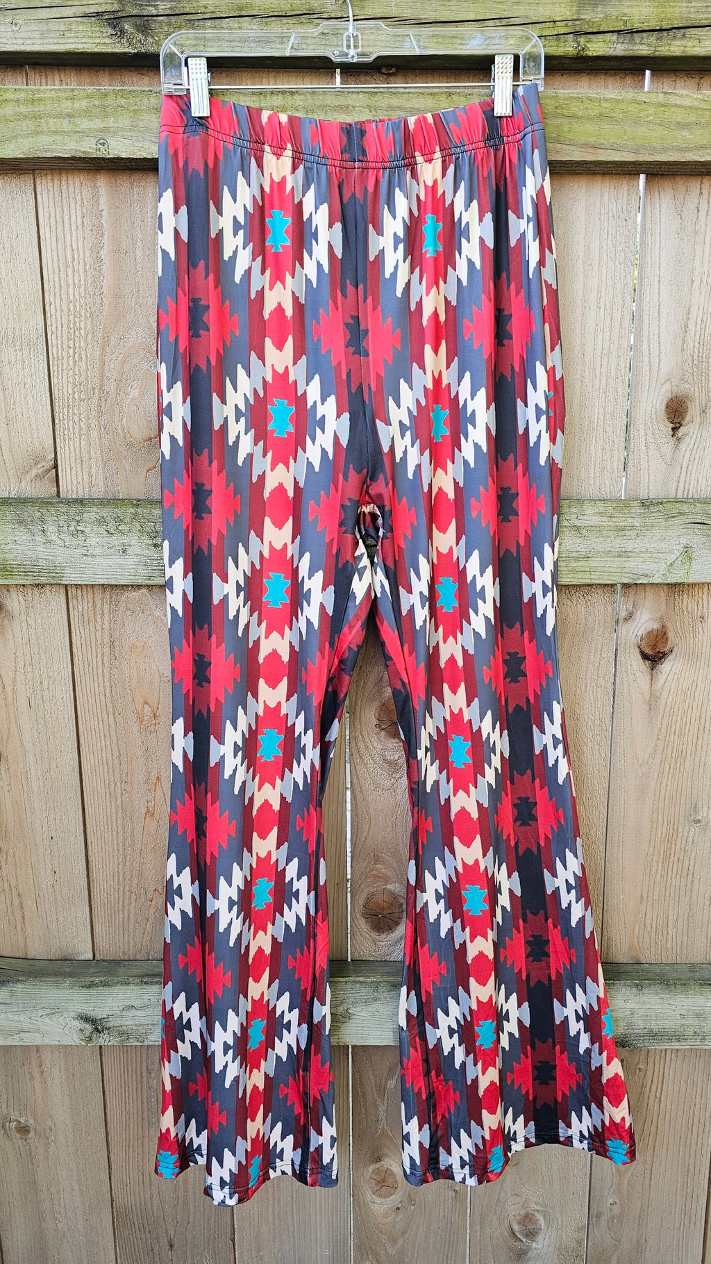 Sedona Aztec Flare Pants showcasing a vibrant southwestern pattern in red, blue, and cream, hanging against a wooden fence.