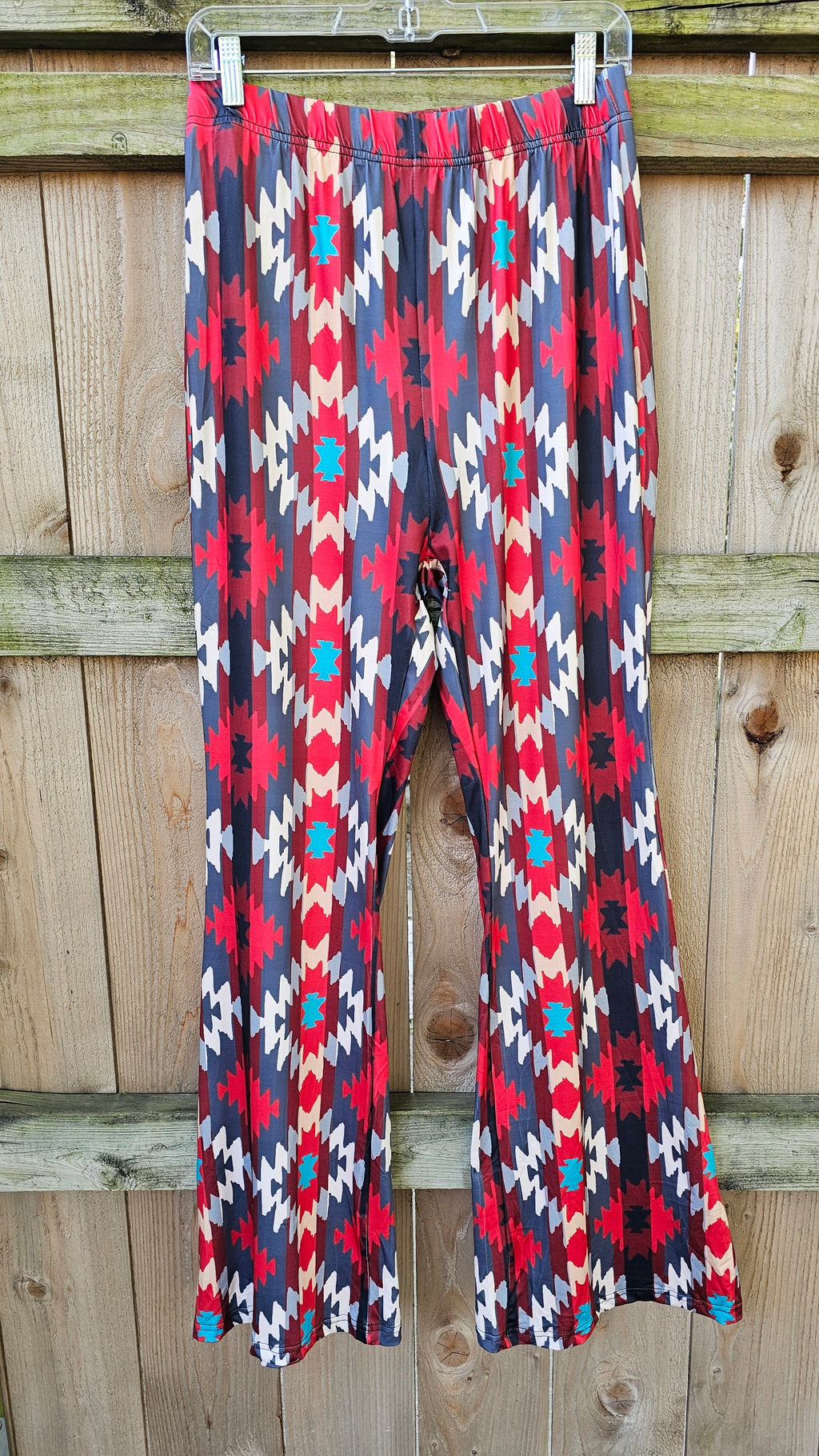 Sedona Aztec Flare Pants with vibrant red, blue, and white pattern hanging on wooden fence.