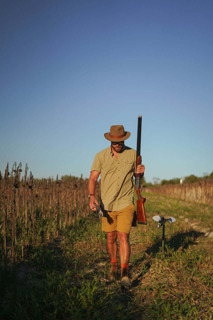 Man in a brimmed field hat walking through a field, carrying a rifle and hunting gear.