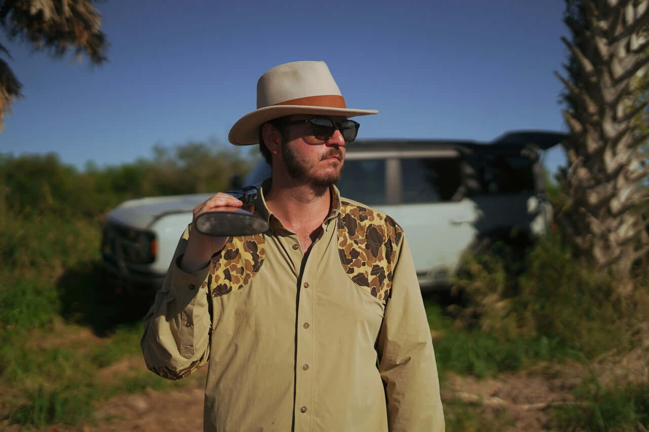 Man wearing a brimmed felted wool field hat and leopard print shirt, holding a beverage outdoors.