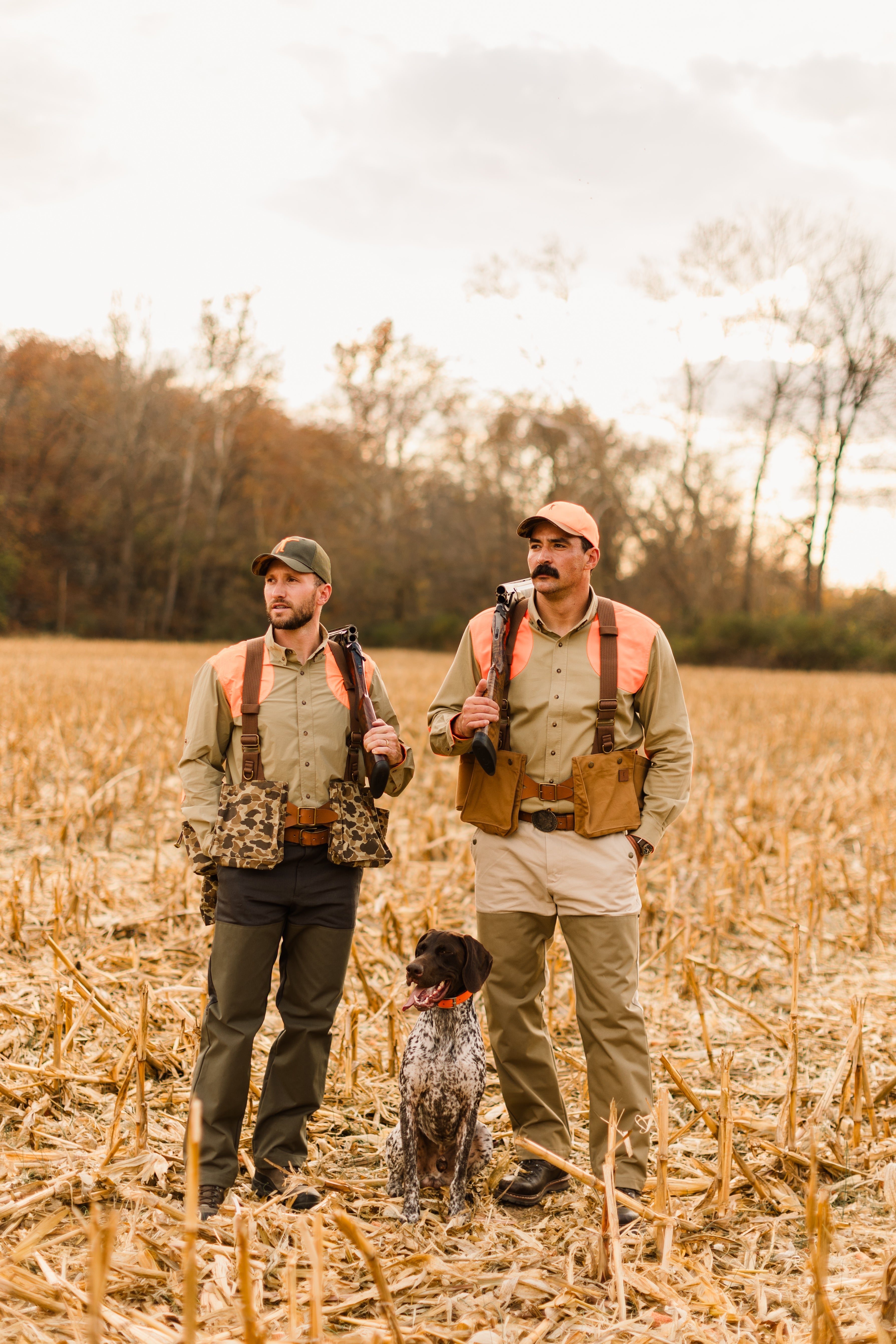 Two hunters wearing outdoor gear stand in a field with a dog, ready for a day in nature.