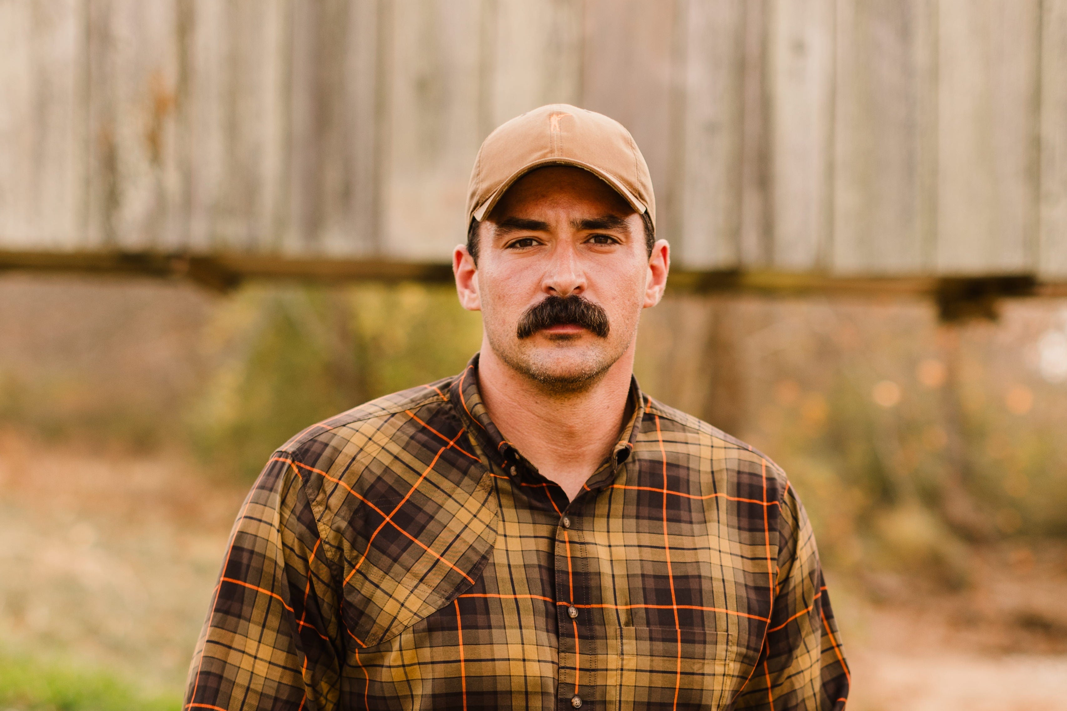 Man wearing a waxed cotton hat and plaid shirt outdoors, showcasing rugged outdoor style.