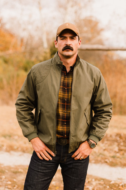 Man wearing an olive green bomber jacket with plaid shirt and waxed cotton hat in an outdoor fall setting.