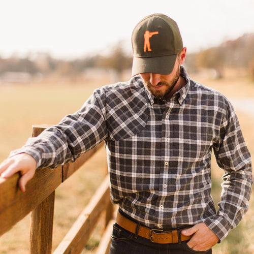 Man wearing Arthur Core Snapback Hat and plaid shirt, standing by a wooden fence in an outdoor setting.