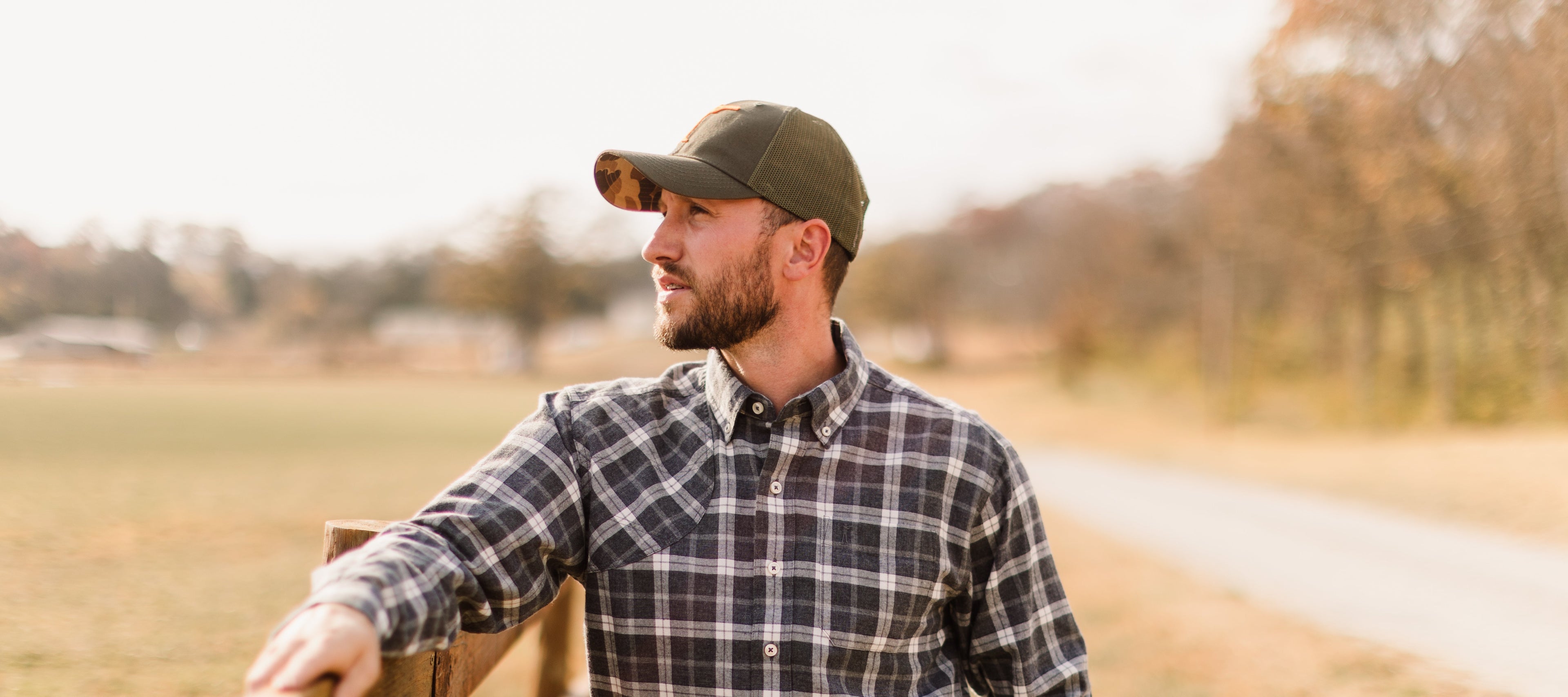 Man wearing Arthur Core Snapback Hat and plaid shirt, standing by a wooden fence in a sunny outdoor setting.