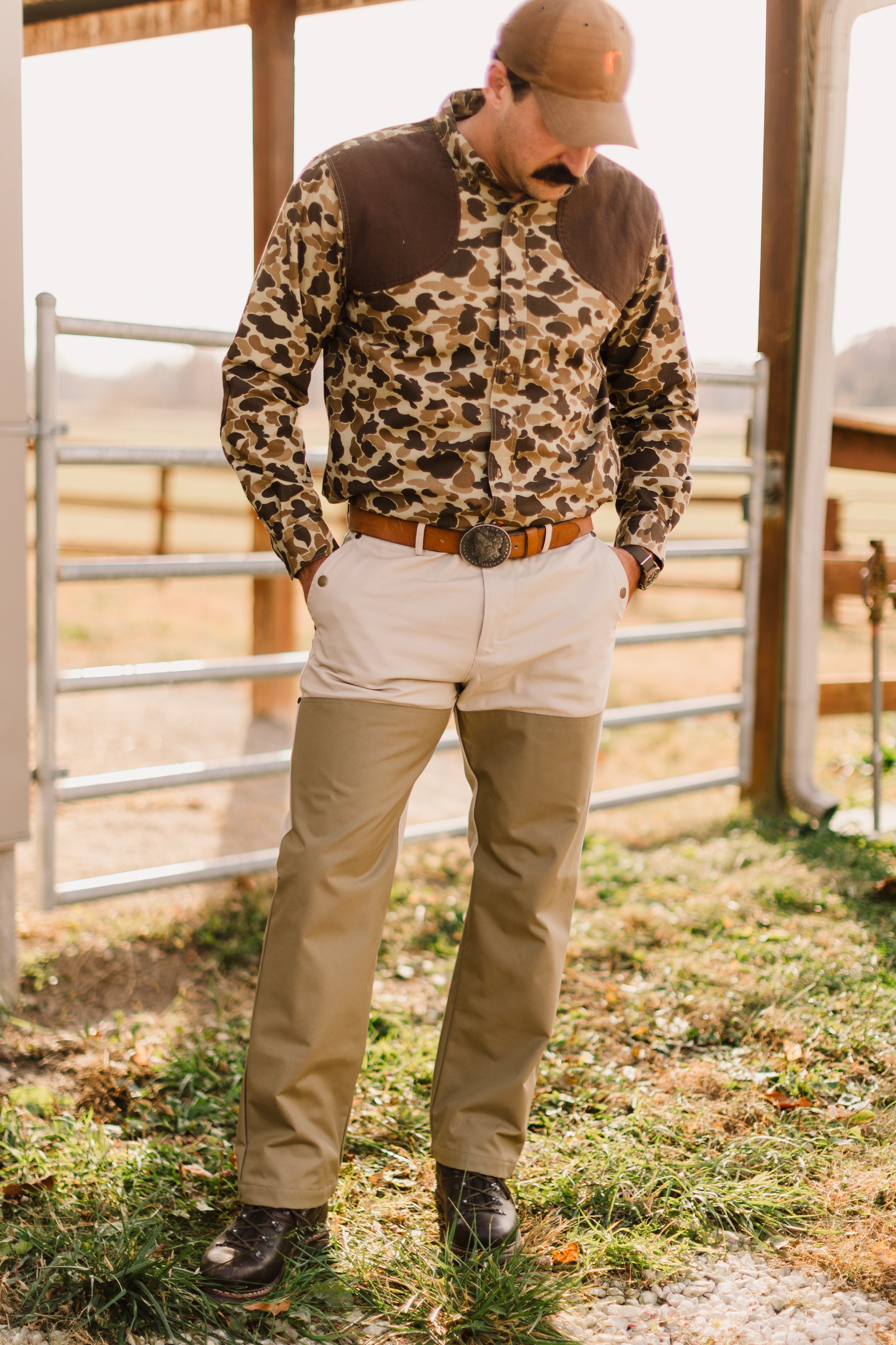 A man wearing a camo pattern shirt and khaki pants stands in a field, showcasing rugged outdoor style.