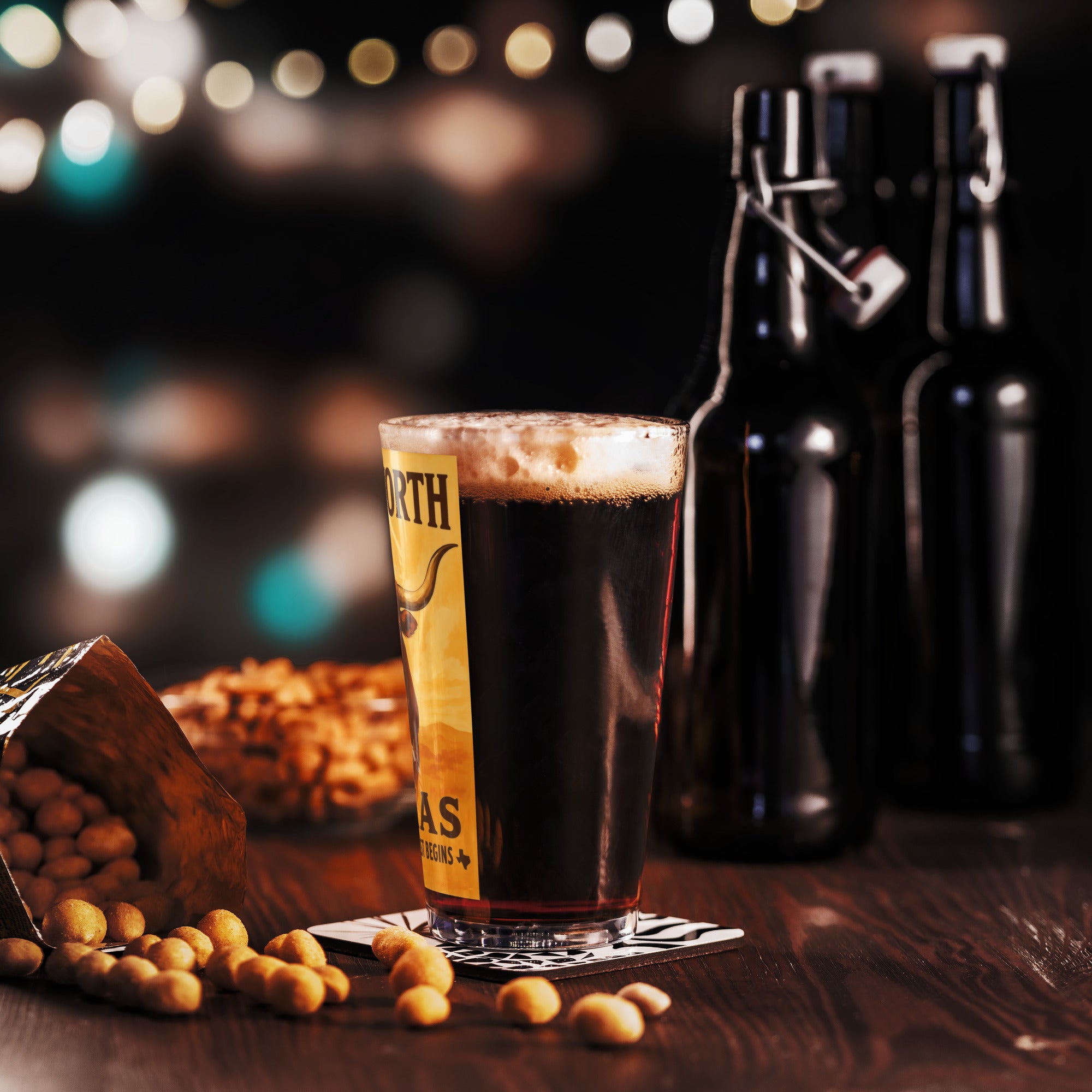 Vintage Fort Worth, Texas pint glass filled with dark beer, surrounded by snacks and bottles on a wooden table.