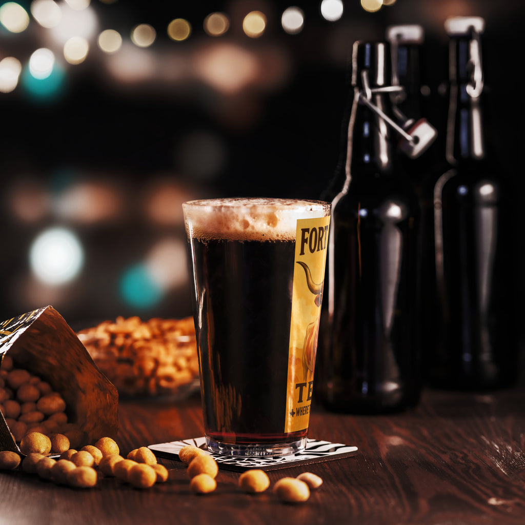 Vintage Fort Worth pint glass with dark beer, peanuts, and beer bottles on a wooden table, glowing bokeh in background.