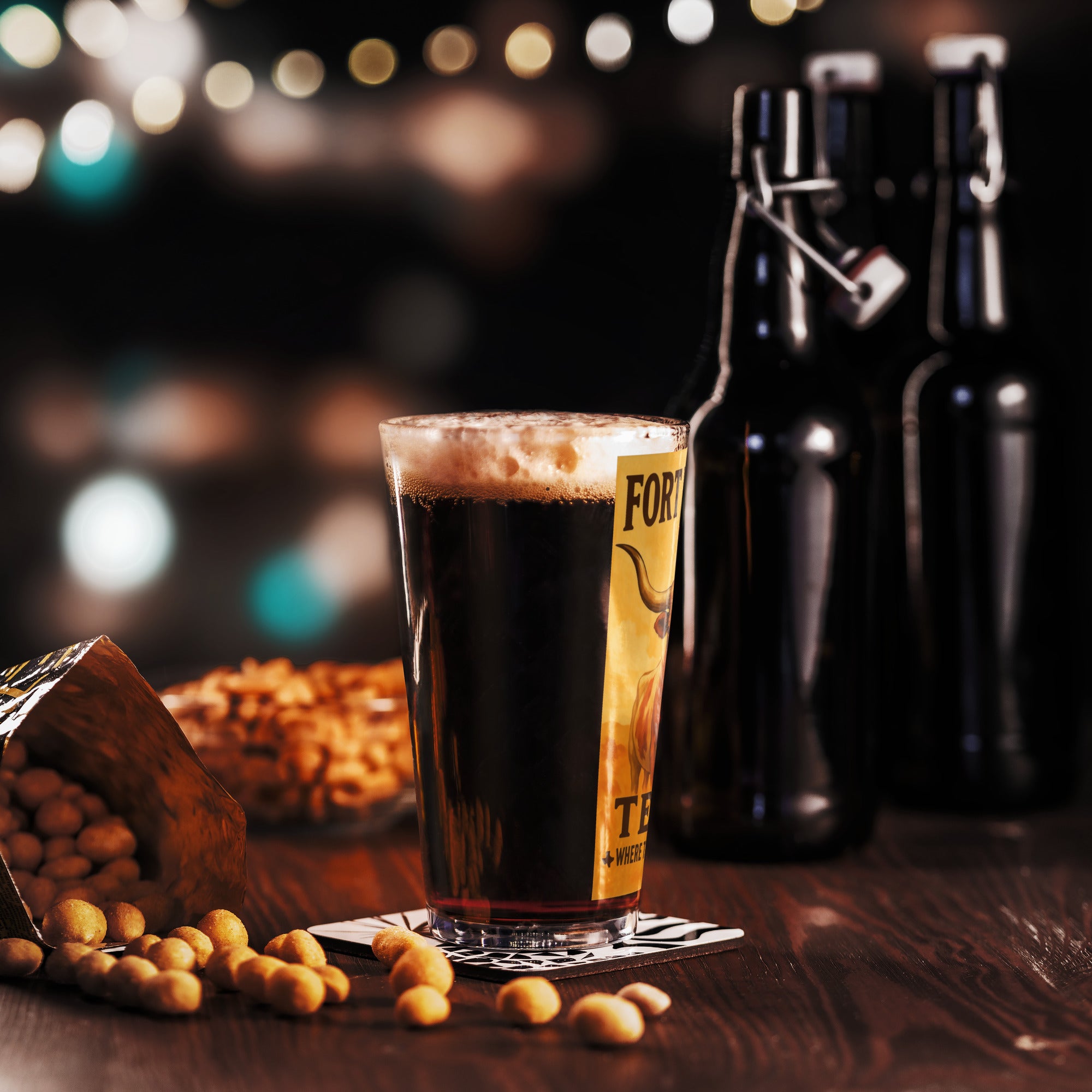 Vintage Fort Worth pint glass with dark beer, peanuts, and beer bottles on a wooden table, glowing bokeh in background.
